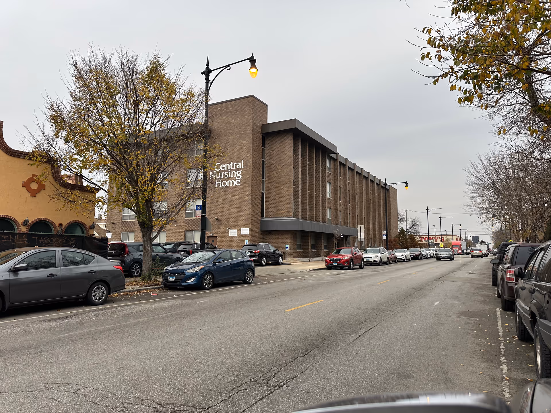 Brick multi-story nursing home building labeled "Central Nursing Home" on a city street with parked cars and leafless trees.