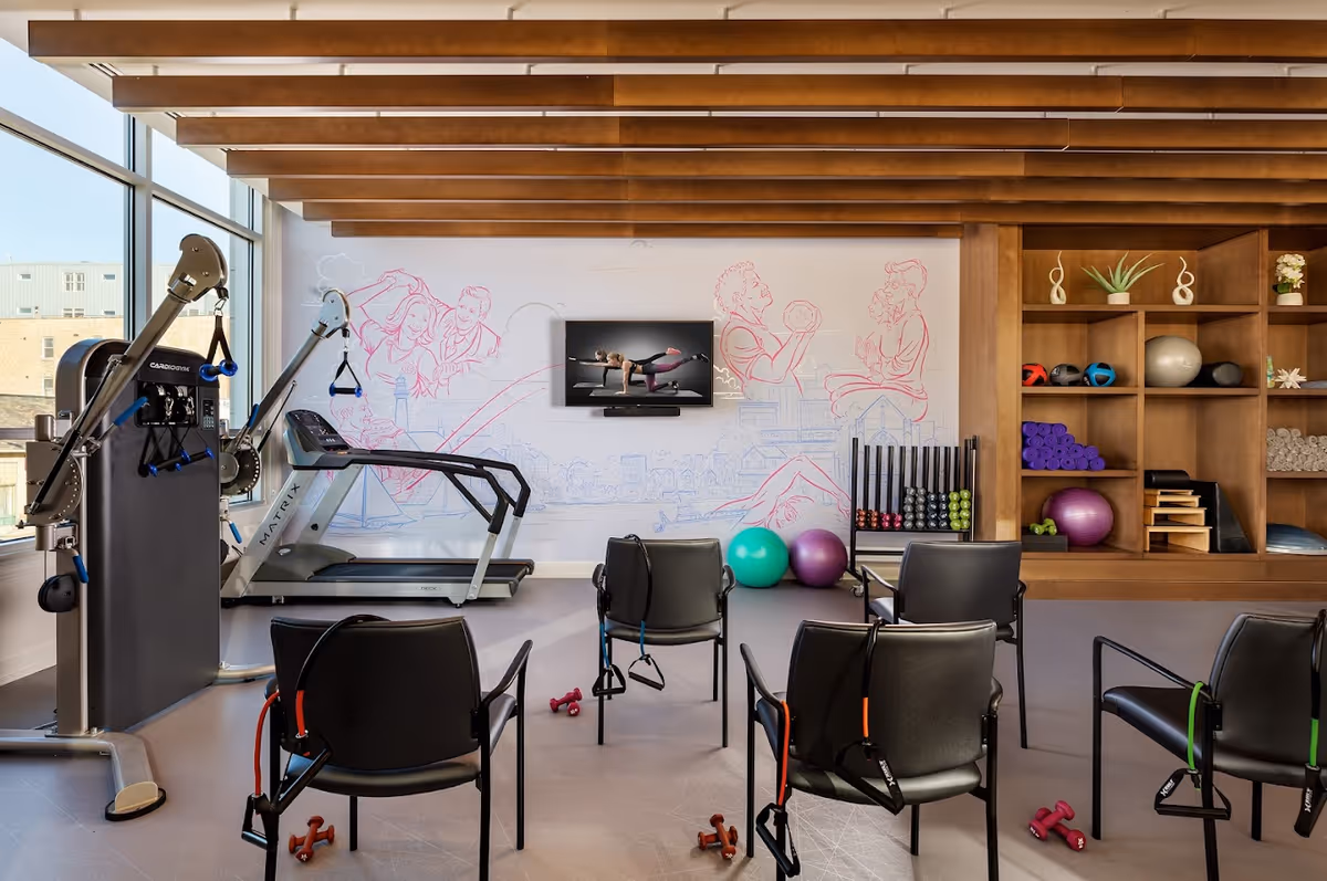 A fitness room with exercise equipment including a treadmill, resistance machines, dumbbells, exercise balls, and chairs equipped with resistance bands. The room has large windows, wooden ceiling beams, and a wall-mounted TV displaying a workout video. Shelves on the right hold various fitness accessories and plants.