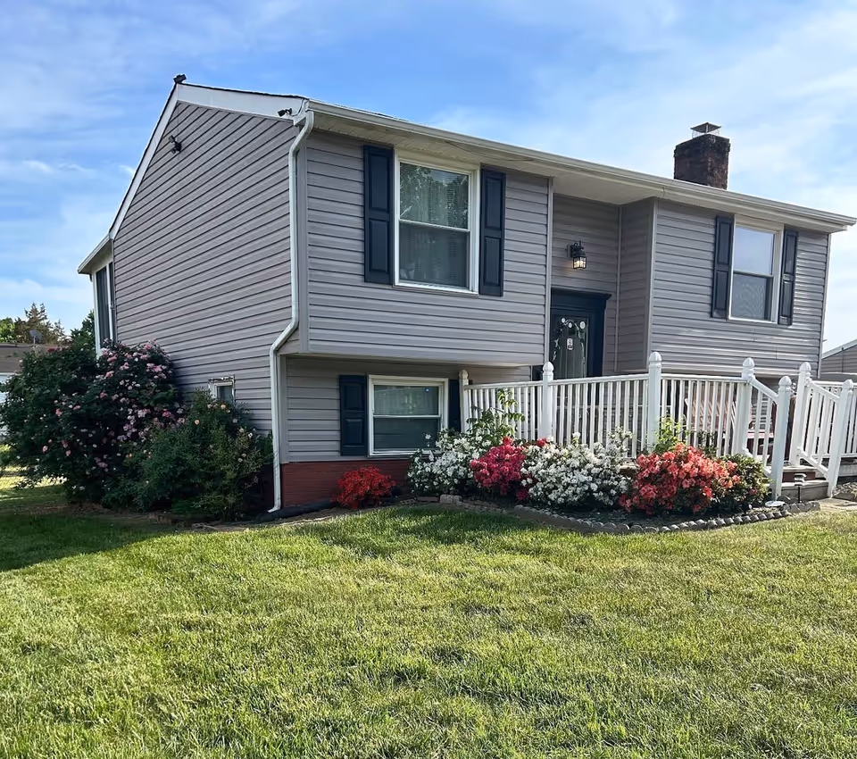 Two-story gray house with white railings and colorful flower beds in a well-kept yard.