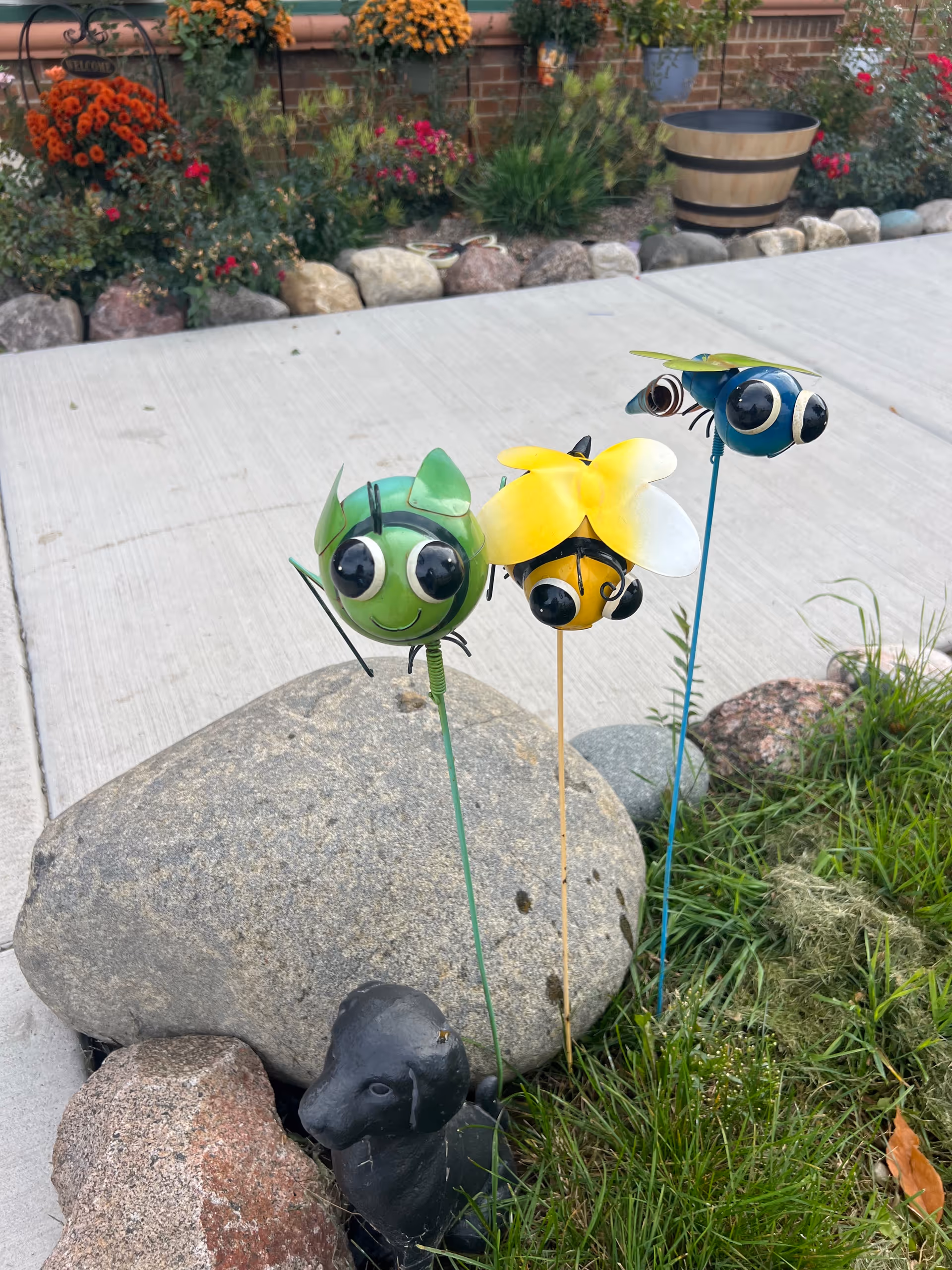 Outdoor garden with colorful flowers and whimsical insect wind spinners beside rocks.