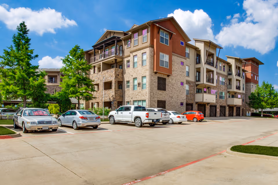 Exterior view of a multi-story senior living facility building with a parking lot in front. Several cars are parked, and there are green trees around the building under a blue sky with some clouds.