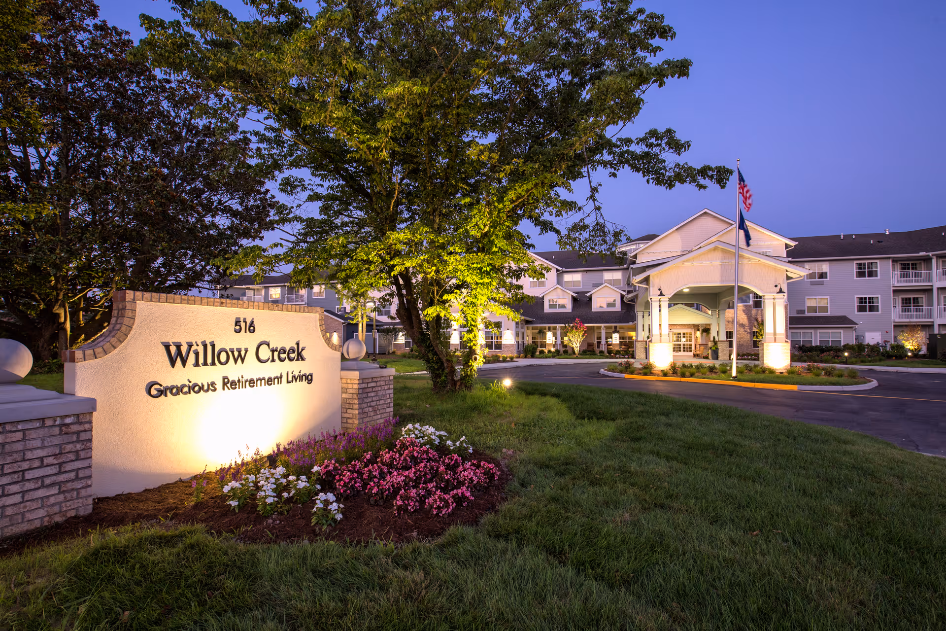 Exterior view of Willow Creek Gracious Retirement Living facility at dusk, showing the illuminated entrance with a covered driveway, an American flag on a flagpole, well-maintained landscaping with green grass, trees, and colorful flowers, and a large sign with the facility name and address.