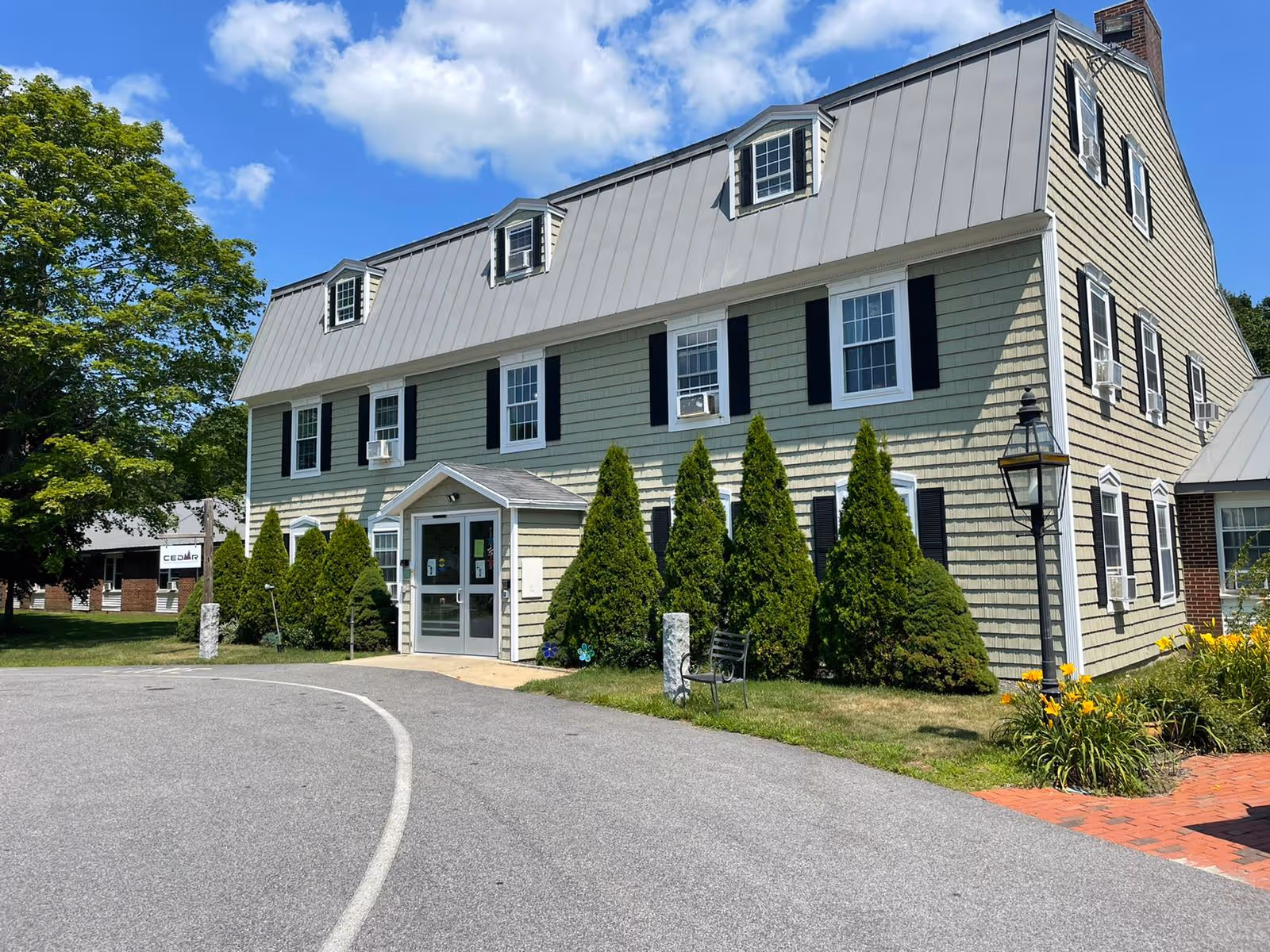 Two-story green senior living building with dormer windows, entrance, and manicured shrubs.