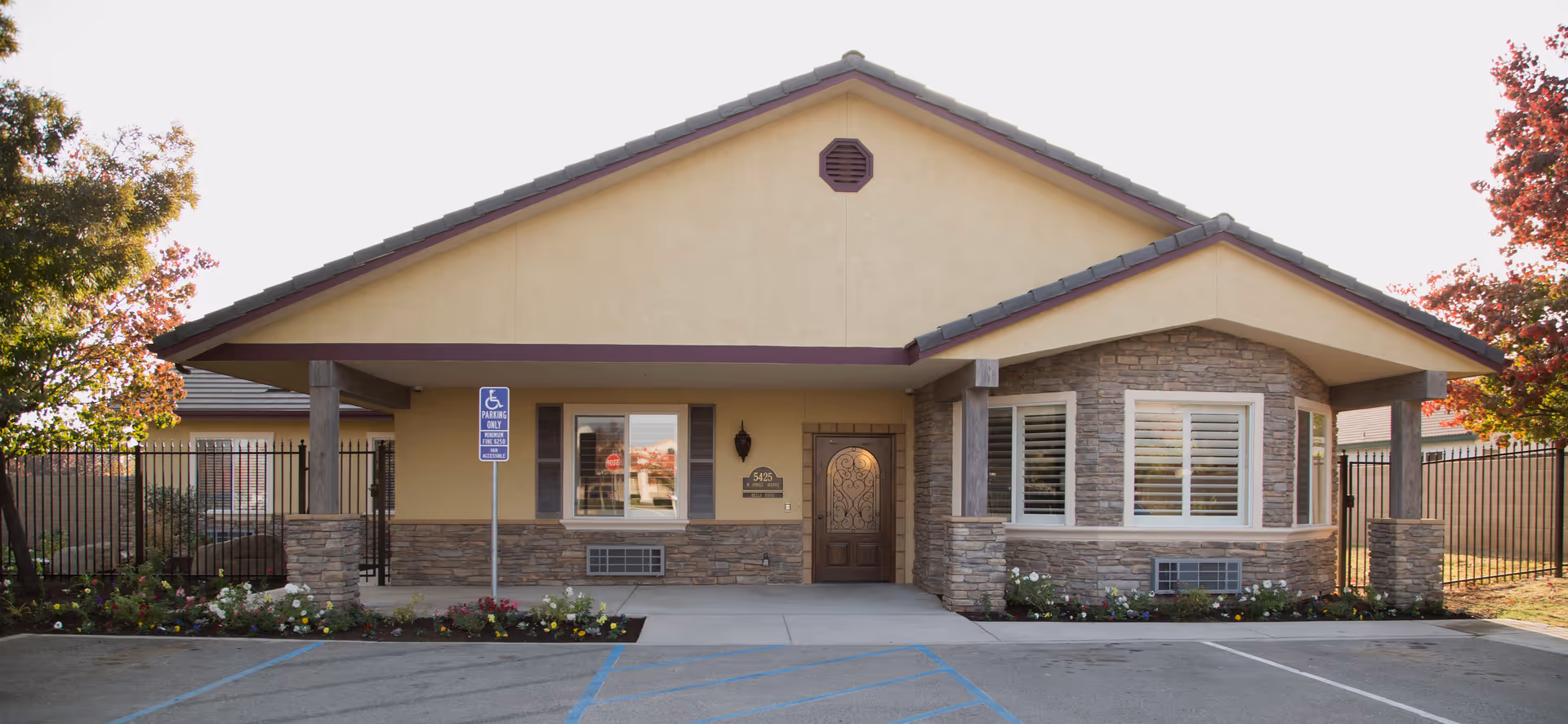 Front exterior view of a single-story building with beige stucco walls and stone accents. The building has a gabled roof with gray tiles and a wooden door with decorative ironwork. There are two windows with white shutters and a handicap parking sign in front. Flower beds with colorful flowers line the front of the building, and trees with autumn foliage are visible on either side.