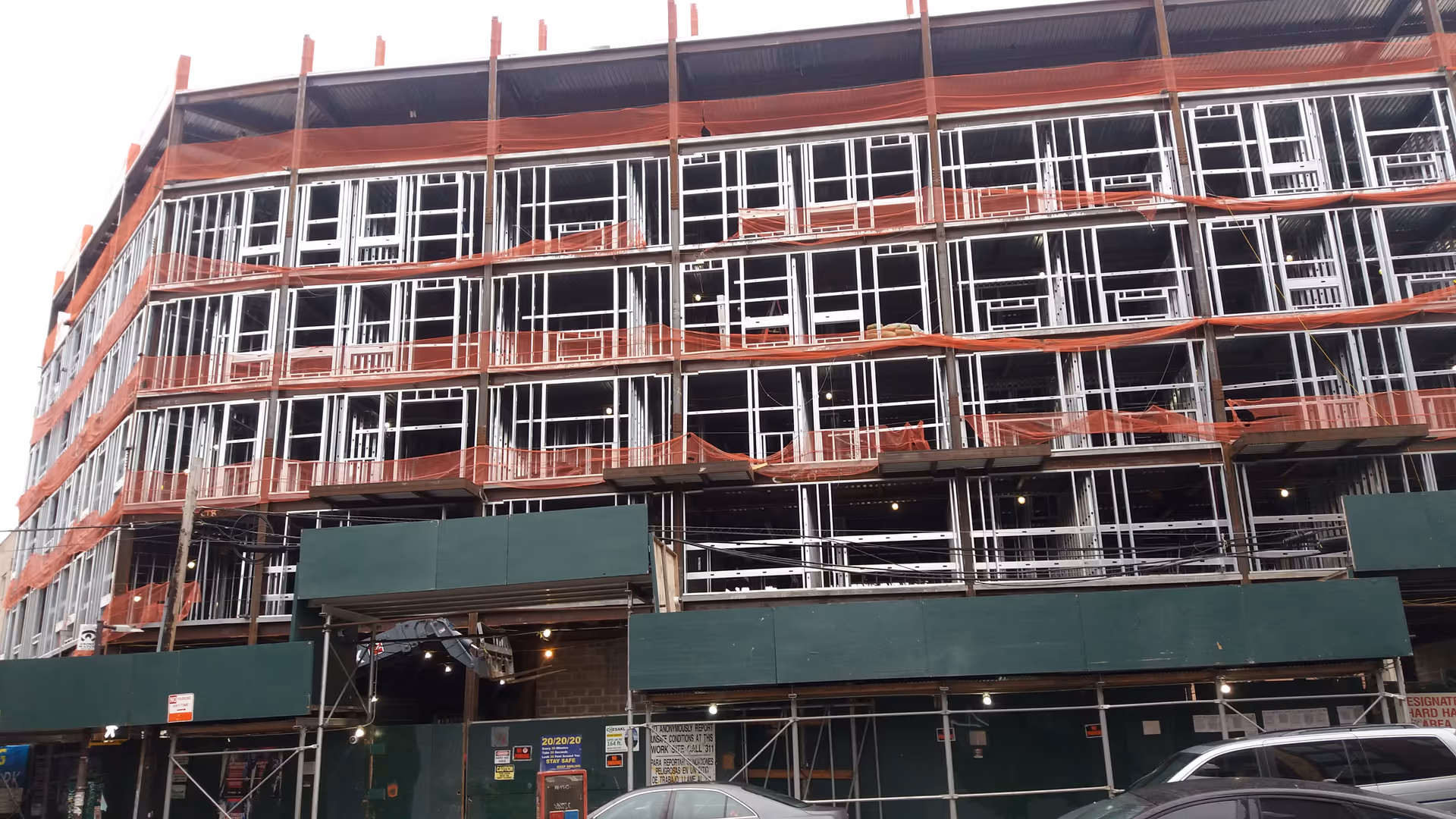 Under construction, multi-story building with metal framing, orange safety netting, and scaffolding.