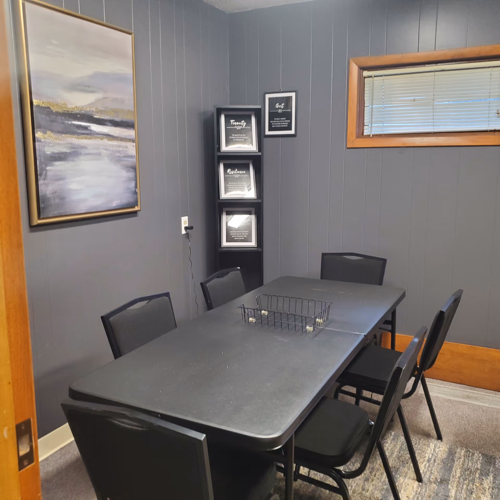A small conference-style room with a gray table, chairs, wall art, and a window.