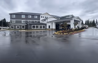 Exterior view of a three-story assisted living facility with a covered entrance and a circular driveway. The building has gray and white siding with multiple windows. The sky is overcast and the ground is wet from recent rain.