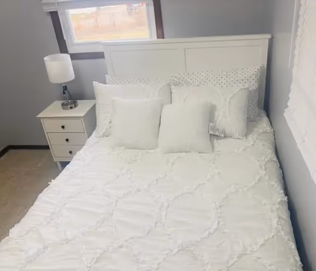 Cozy white bedroom featuring a bed, nightstand, lamp, and a window.