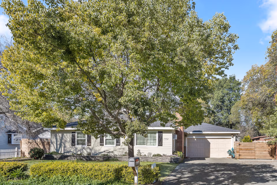 Front exterior of a single-story house with a large leafy tree, driveway, garage, and mailbox.