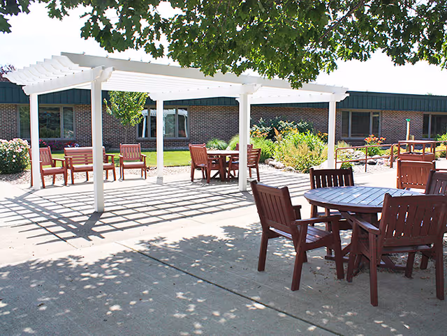 Outdoor patio with wooden tables and chairs under a white pergola at a senior living campus.