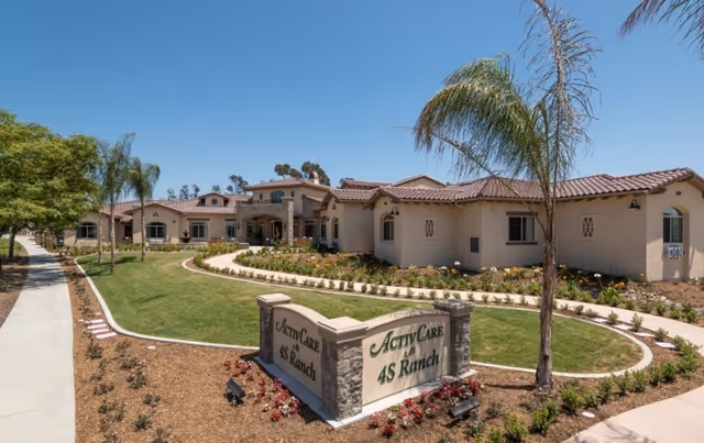 Exterior view of ActivCare 4S Ranch facility showing a single-story building with a tiled roof, surrounded by landscaped gardens and palm trees under a clear blue sky. A sidewalk curves around the lawn leading to the entrance, and a stone sign with the facility name is prominently displayed in the foreground.