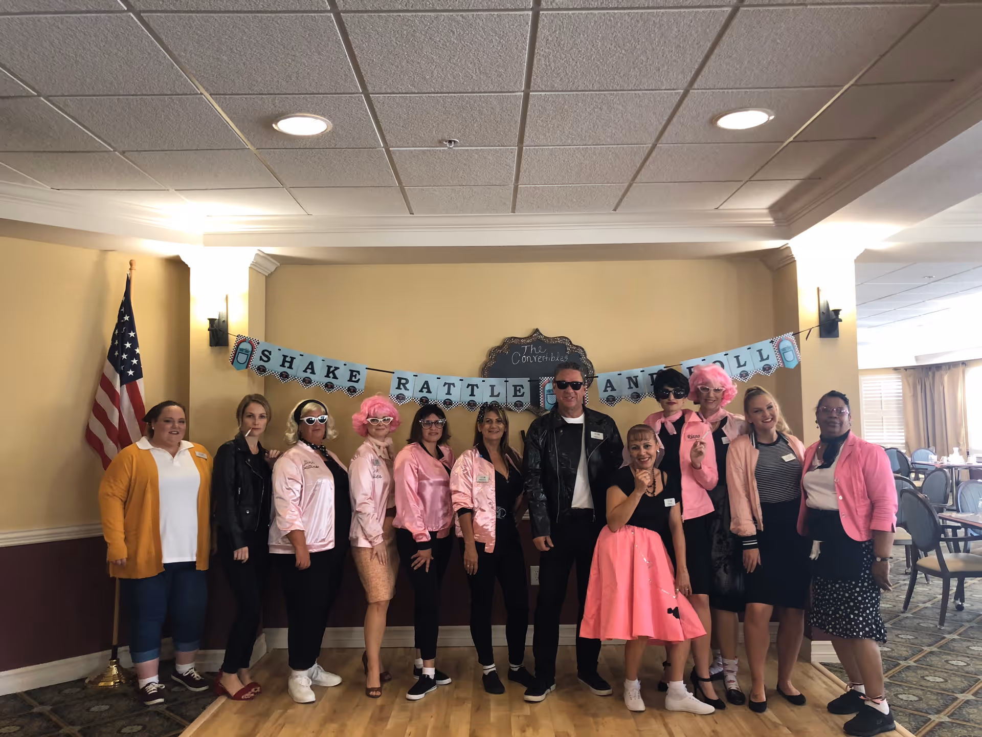 A group of eleven adults standing indoors in front of a beige wall with a banner that reads 'Shake Rattle and Roll'. Most people are dressed in 1950s-style clothing, including pink jackets, sunglasses, and retro hairstyles. An American flag is visible on the left side, and a dining area with tables and chairs is visible on the right side.