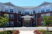Two-story brick senior living facility with central entrance, glass doors, and manicured landscaping.