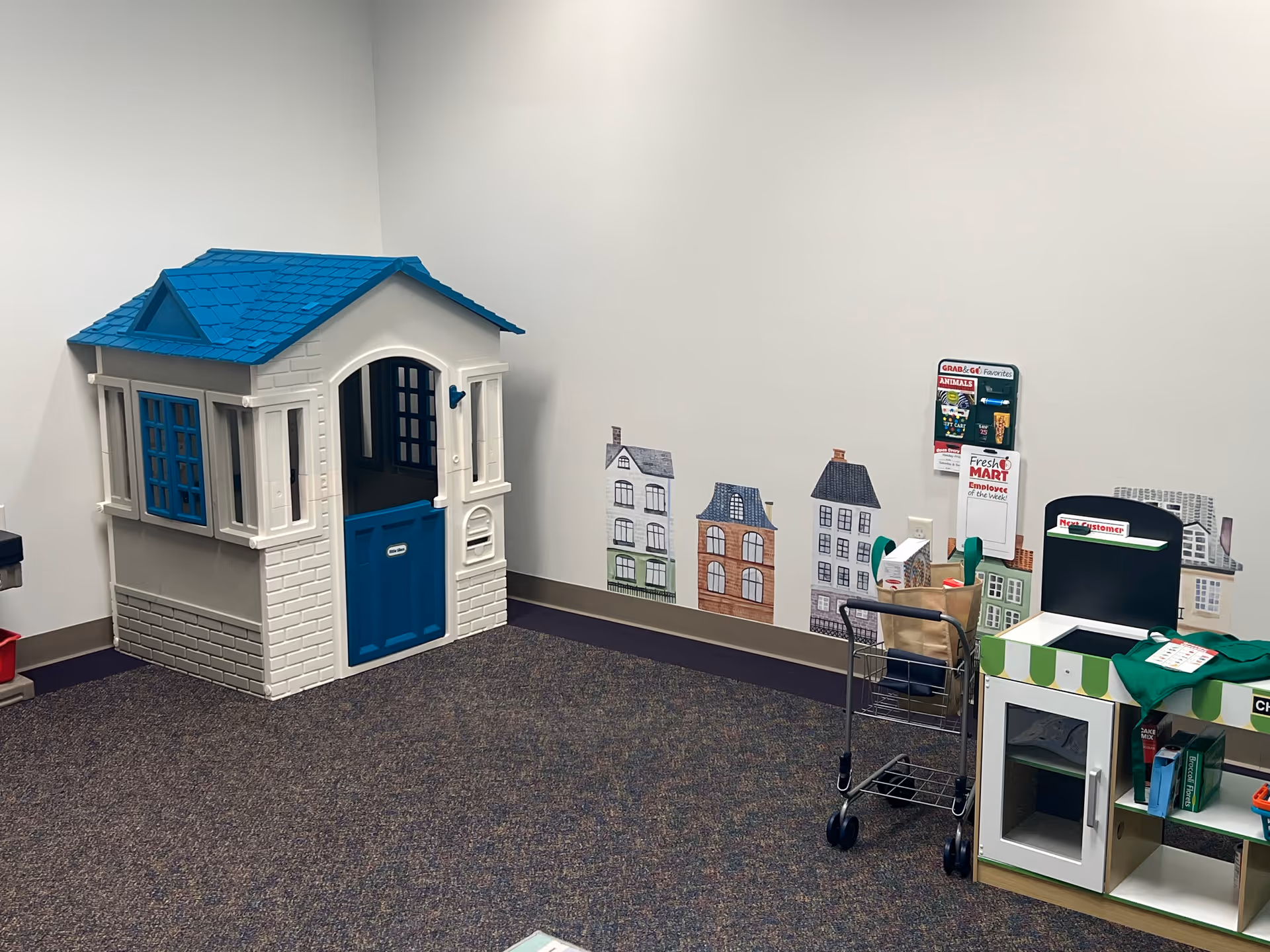 Indoor play area with a blue-roofed toy house, colorful building decals, and a cart.
