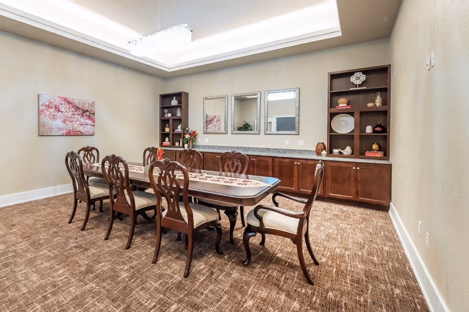 A dining room with a long wooden table surrounded by eight matching chairs with cushioned seats. The room features a beige carpet, light-colored walls, and a ceiling with recessed lighting and a modern chandelier. On one wall, there are two built-in wooden cabinets with shelves displaying decorative items, and three mirrors hang above the cabinets. A floral painting is on the adjacent wall.