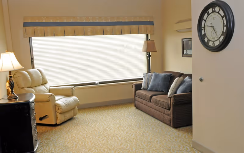 Cozy living room with beige furniture, large window blinds, and a wall clock.