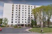 White multi-story senior living building with trees, parking lot, and curved driveway.