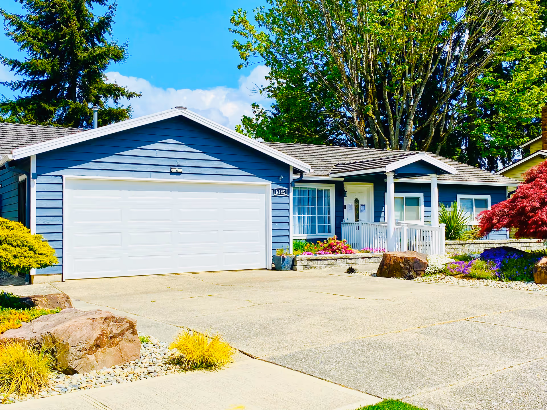 Blue single-story house with white garage doors, front porch, and landscaped yard under bright sunlight.