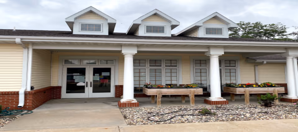 Front view of a yellow senior living facility with columns, windows, and flower planters.
