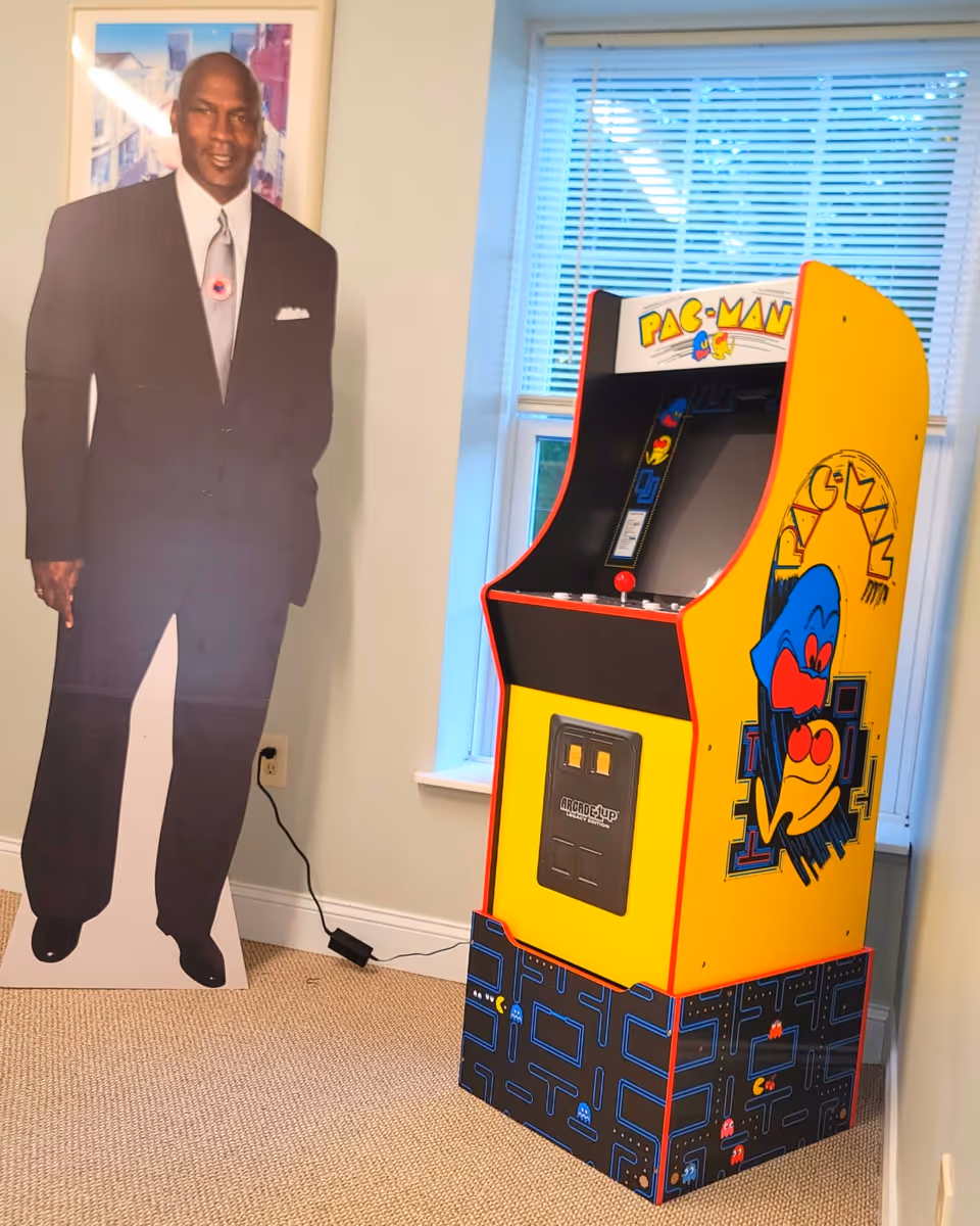 Life-size cardboard cutout of a man beside a Pac-Man arcade cabinet in a room.