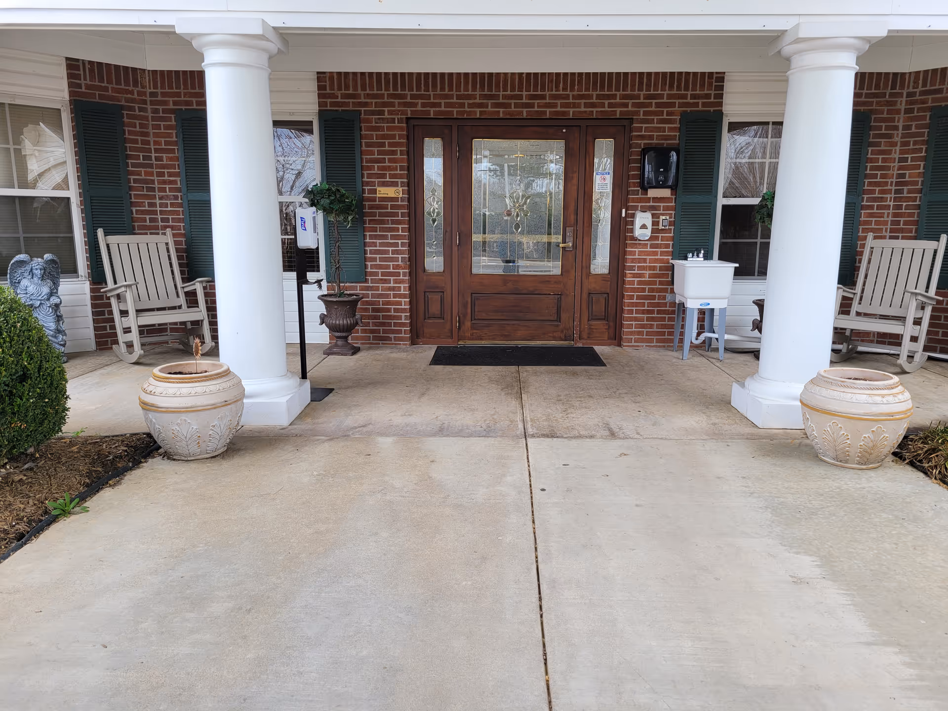 Entrance of a brick rehabilitation and healthcare building with double wooden doors flanked by white columns, rocking chairs, and large urn planters.