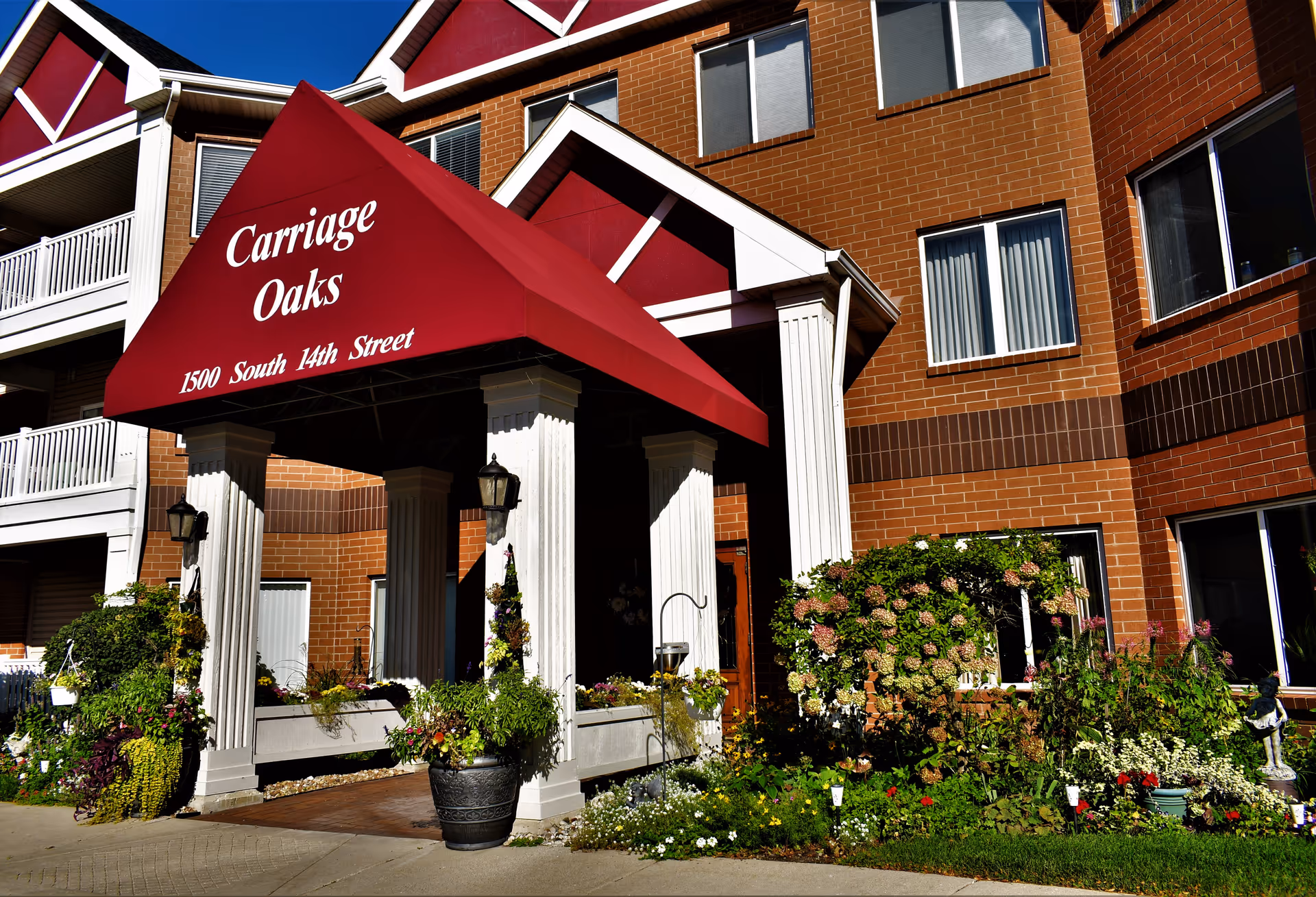 Carriage Oaks senior living entrance with red canopy, brick facade, and flowering garden.