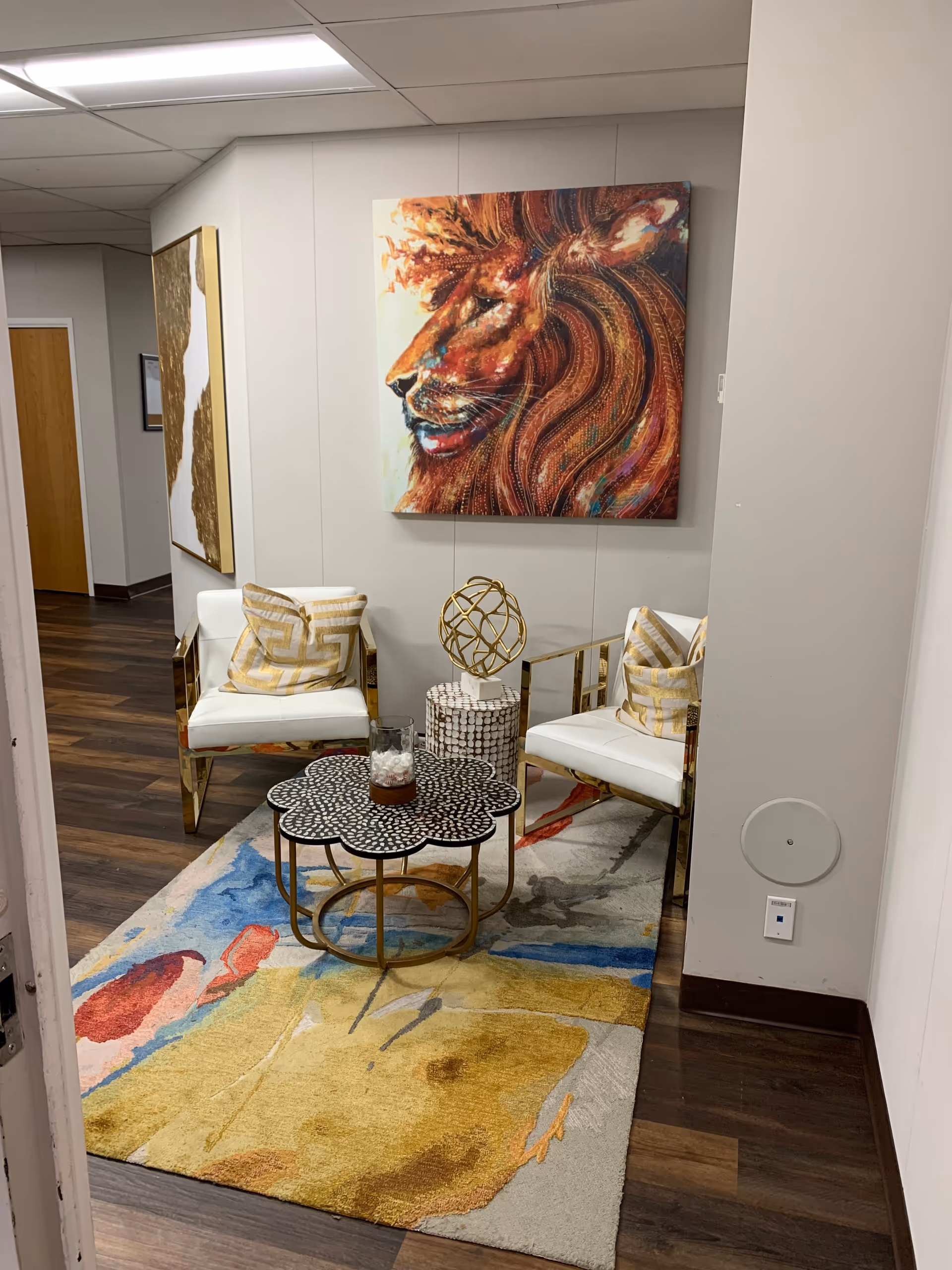 A small modern lounge area with two white-gold chairs, colorful rug, and lion artwork.