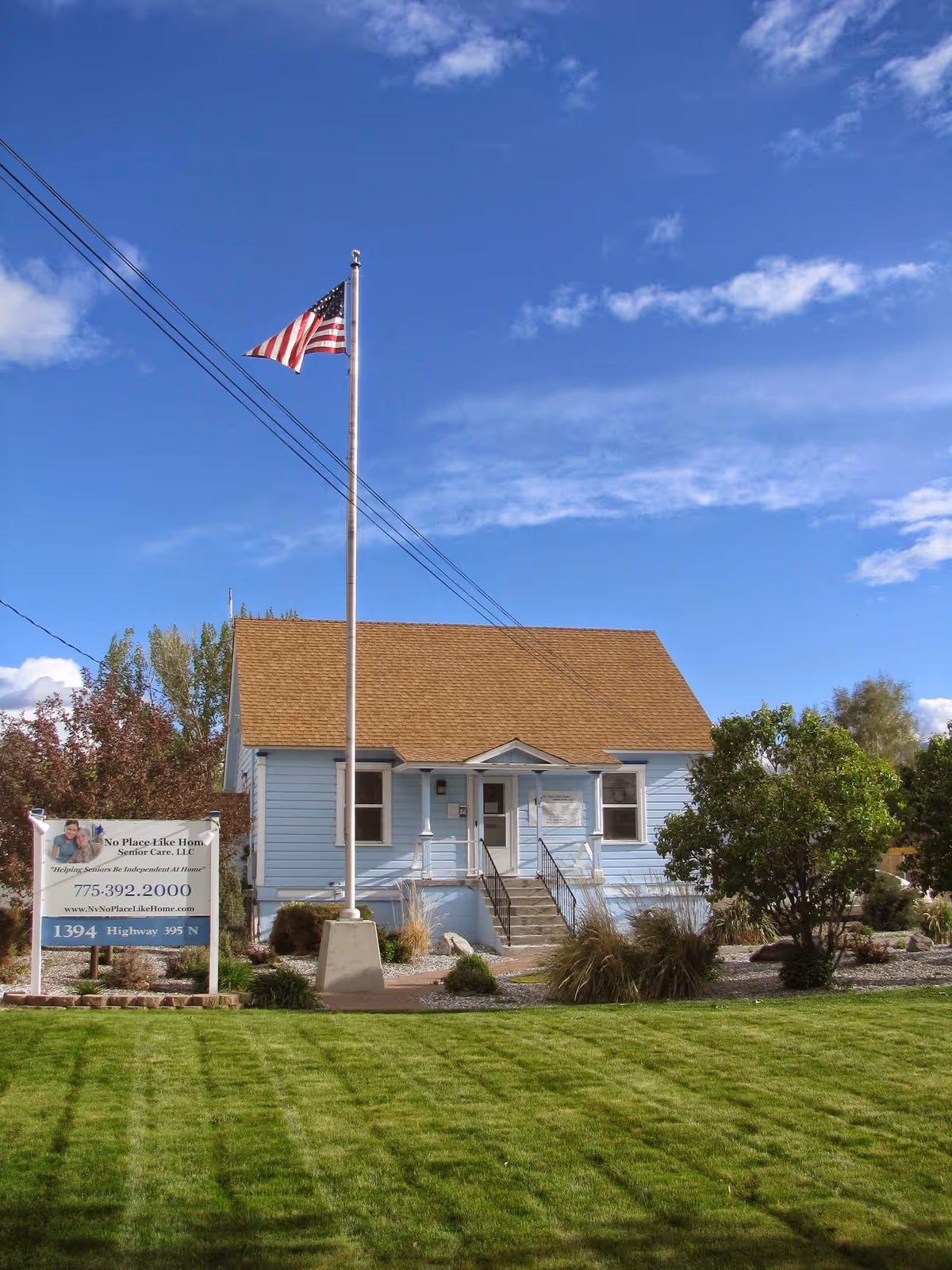 Small light blue senior care building with a flagpole, sign, and manicured lawn.