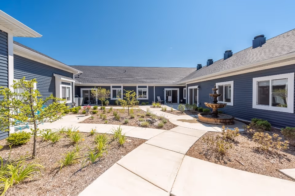 Outdoor courtyard area of a senior living facility with a concrete walkway, small trees, plants, and a multi-tiered water fountain. The building surrounding the courtyard has blue siding and white trim with several windows and doors leading to the courtyard. The sky is clear and blue.