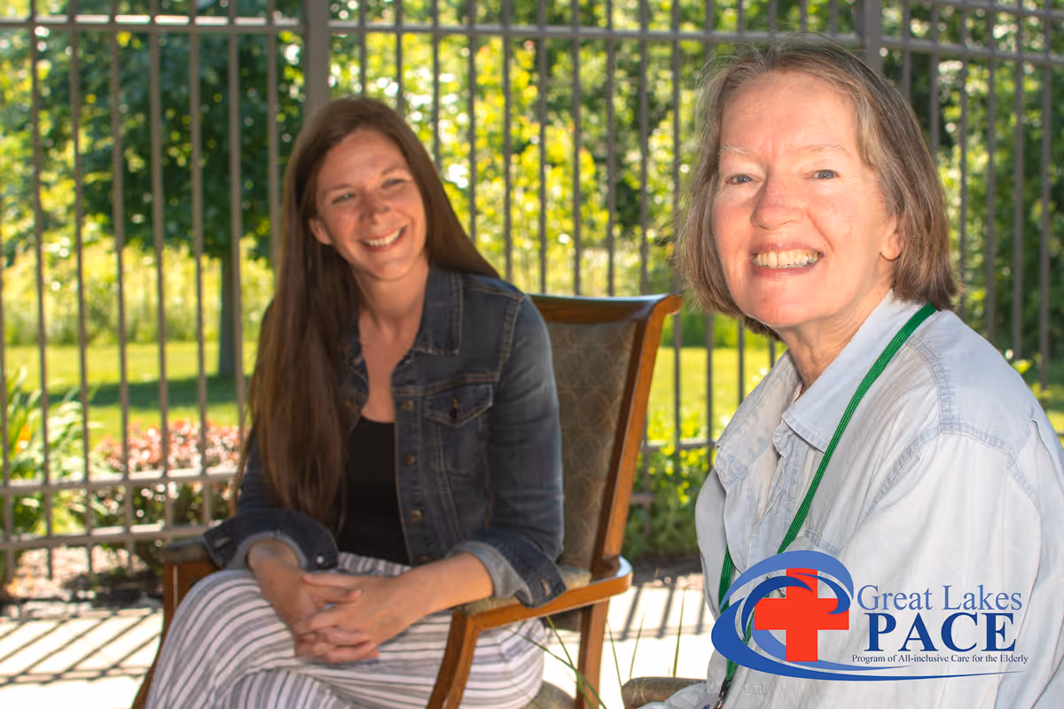 Two smiling women sit outdoors on chairs near a railing, greenery behind, with Great Lakes PACE.