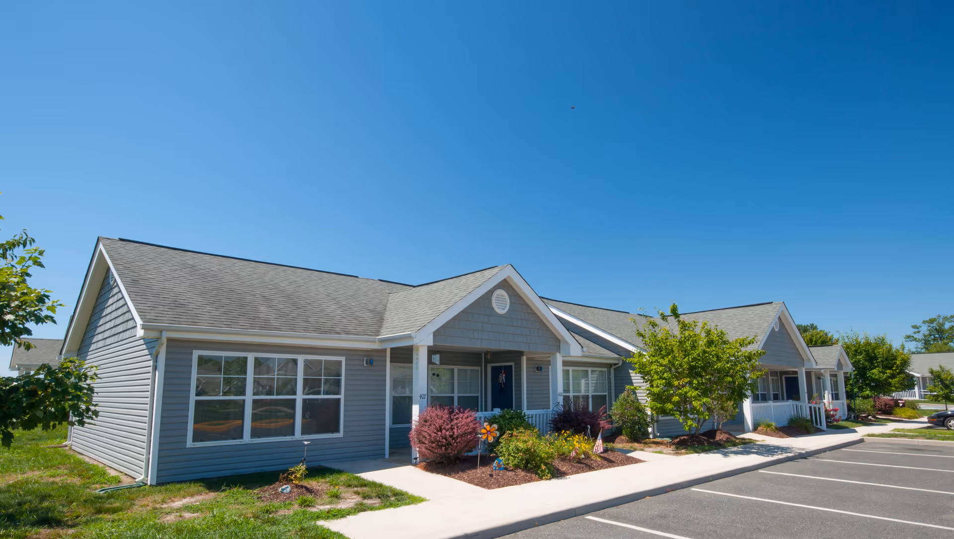 Single-story gray cottages with porches, landscaped front yards and adjacent parking under a clear blue sky.