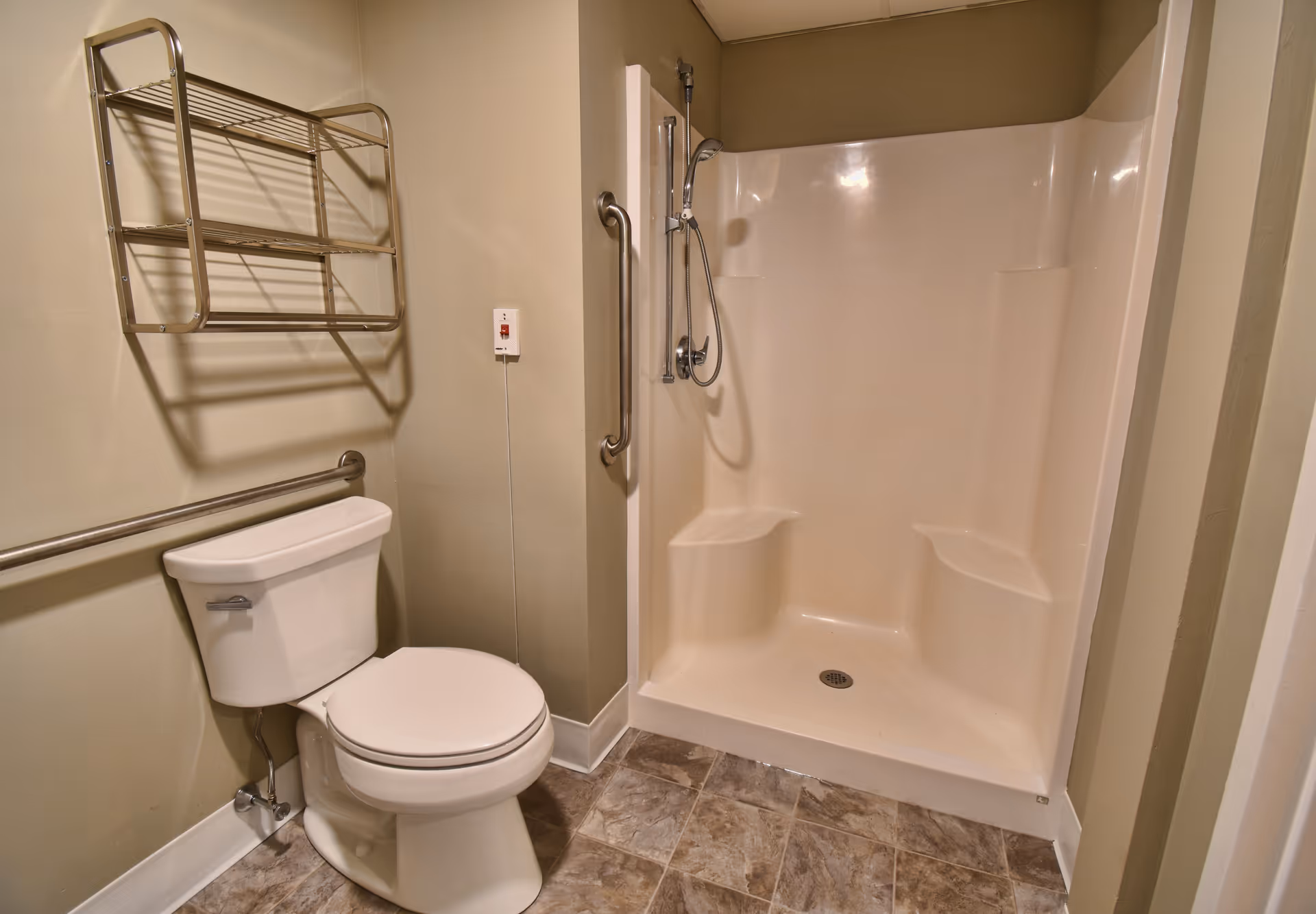 A bathroom with a white toilet, a wall-mounted metal towel rack above it, and a beige walk-in shower with built-in seats and a handheld showerhead. The floor has brown and beige tiles, and there are grab bars installed near the toilet and inside the shower for accessibility.
