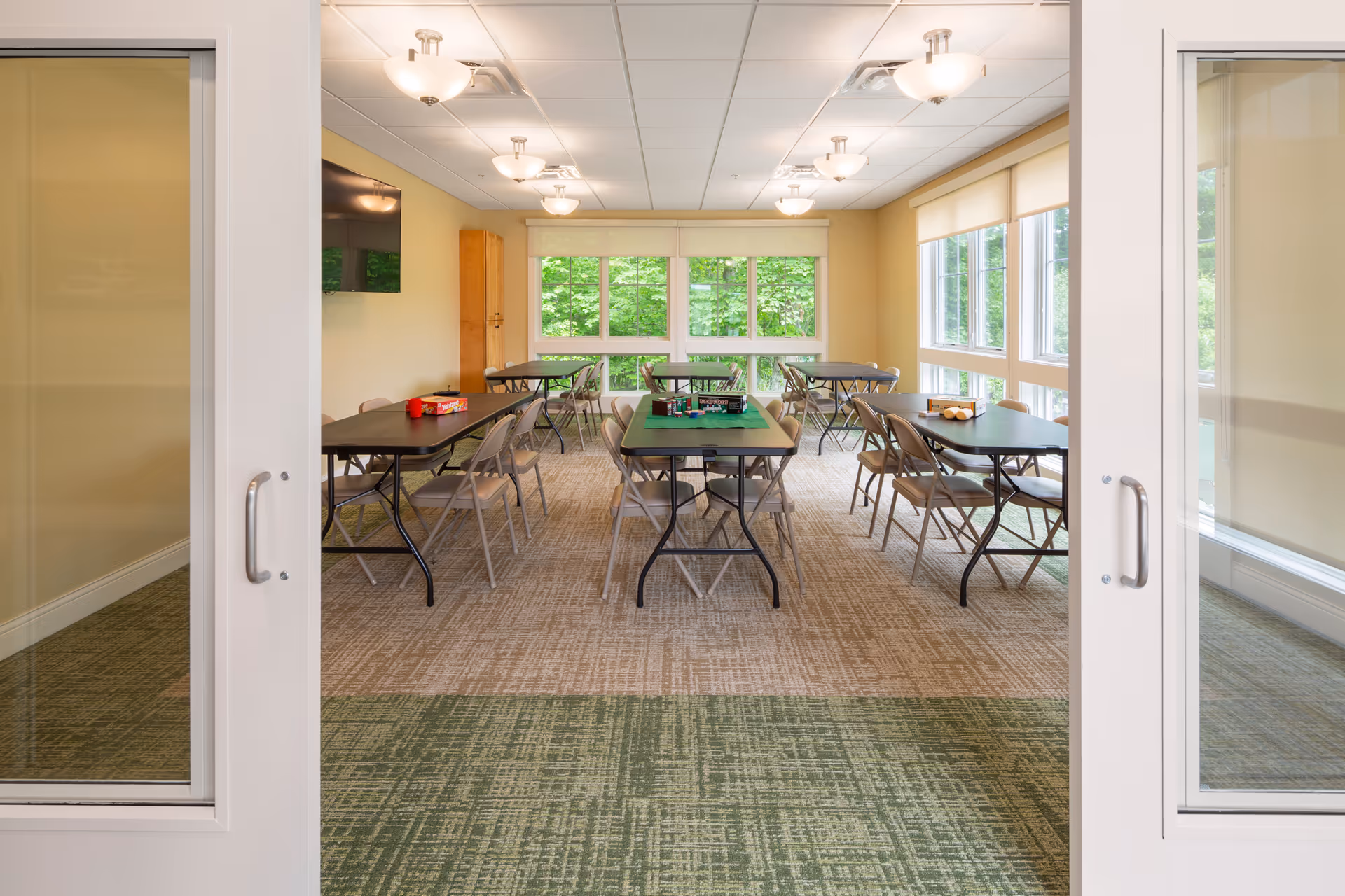 Bright communal dining room with multiple tables, chairs, and large windows in a senior living facility.
