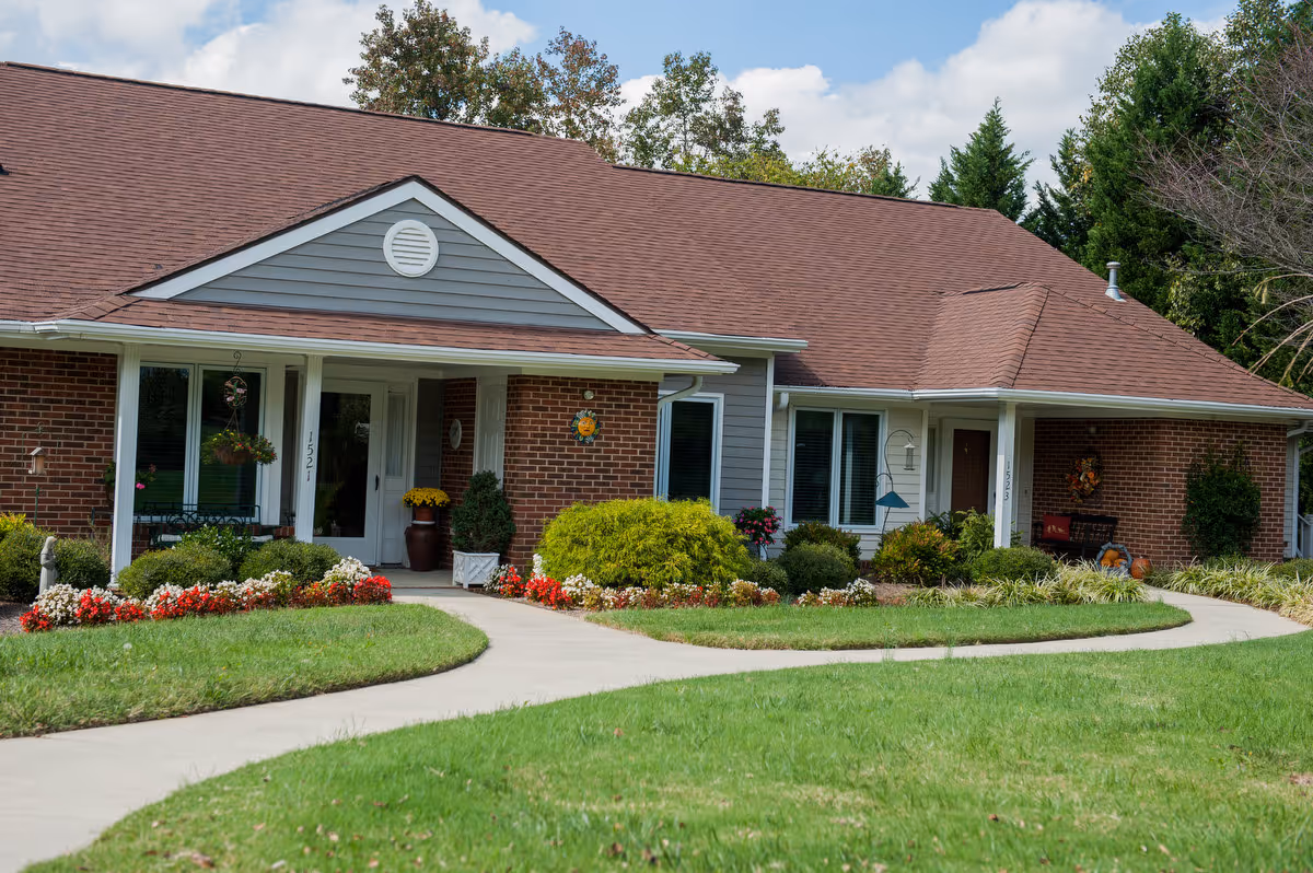 Front of a single-story brick and siding senior living building with a covered porch, flowerbeds, and a curved sidewalk.