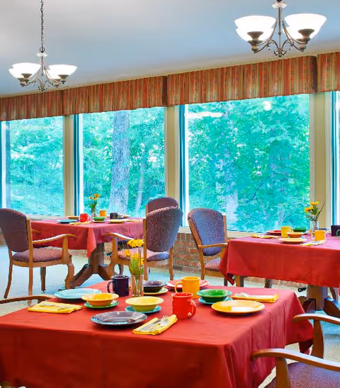 Dining area with several tables covered in red tablecloths, set with colorful plates, cups, and yellow napkins. Chairs with wooden arms surround the tables. Large windows in the background show green trees outside, and two ceiling light fixtures illuminate the room.