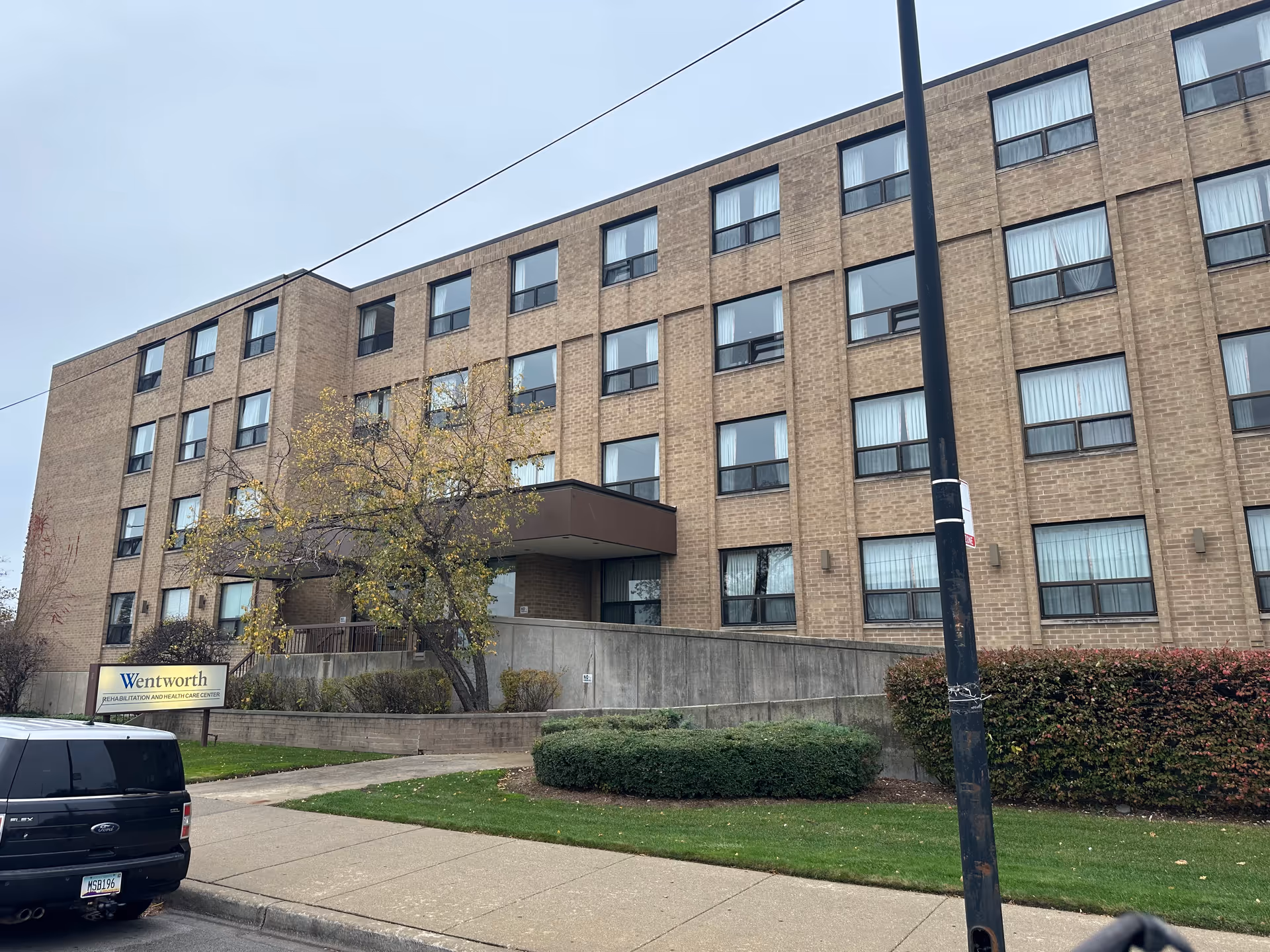 Exterior view of a multi-story brick building with many windows, a small entrance canopy, and a sign in front that reads Wentworth Rehabilitation and Health Care Center. There is a sidewalk, some bushes, a tree with autumn leaves, and a parked black vehicle in front of the building.