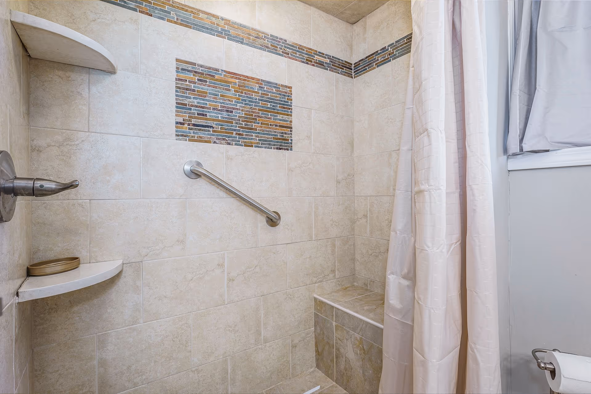Accessible bathroom shower with grab bar, bench, mosaic tile accent, and curtain.