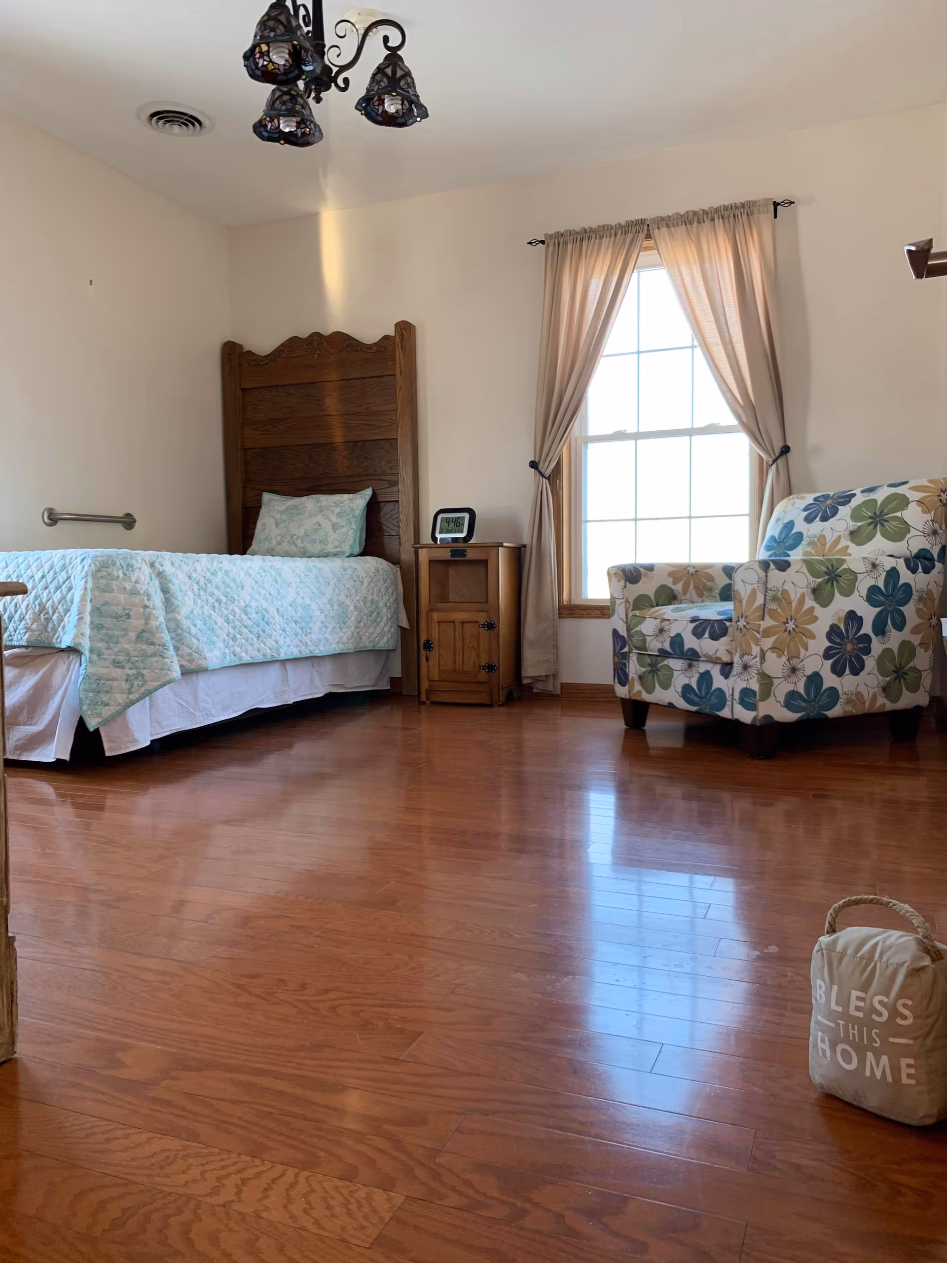 A bright bedroom with wooden flooring, a single bed with a wooden headboard and light blue patterned bedding, a wooden nightstand with a digital clock, a window with beige curtains, and a floral patterned armchair. A small fabric doorstop with the words 'BLESS THIS HOME' is on the floor near the armchair.