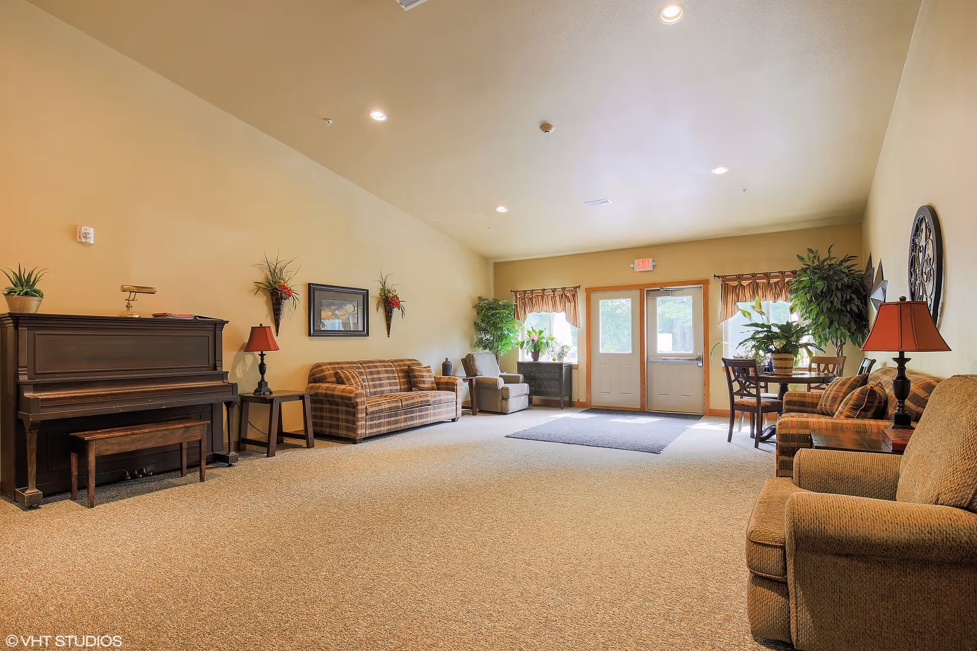 A spacious living room area with beige walls and carpeted floor. The room features a dark wooden piano with a bench, two plaid sofas, an armchair, side tables with lamps, wall-mounted floral decorations, and framed artwork. Near the back, there is a small dining table with chairs and several potted plants by the windows and door letting in natural light.