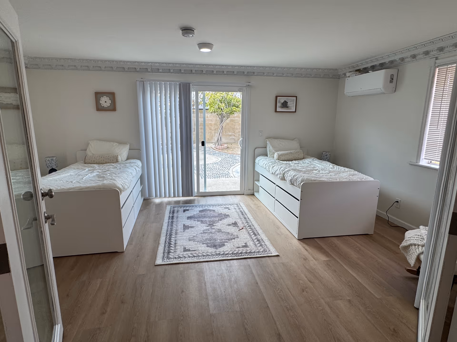 Two twin beds with white storage under, a central rug, and sliding door to a courtyard.