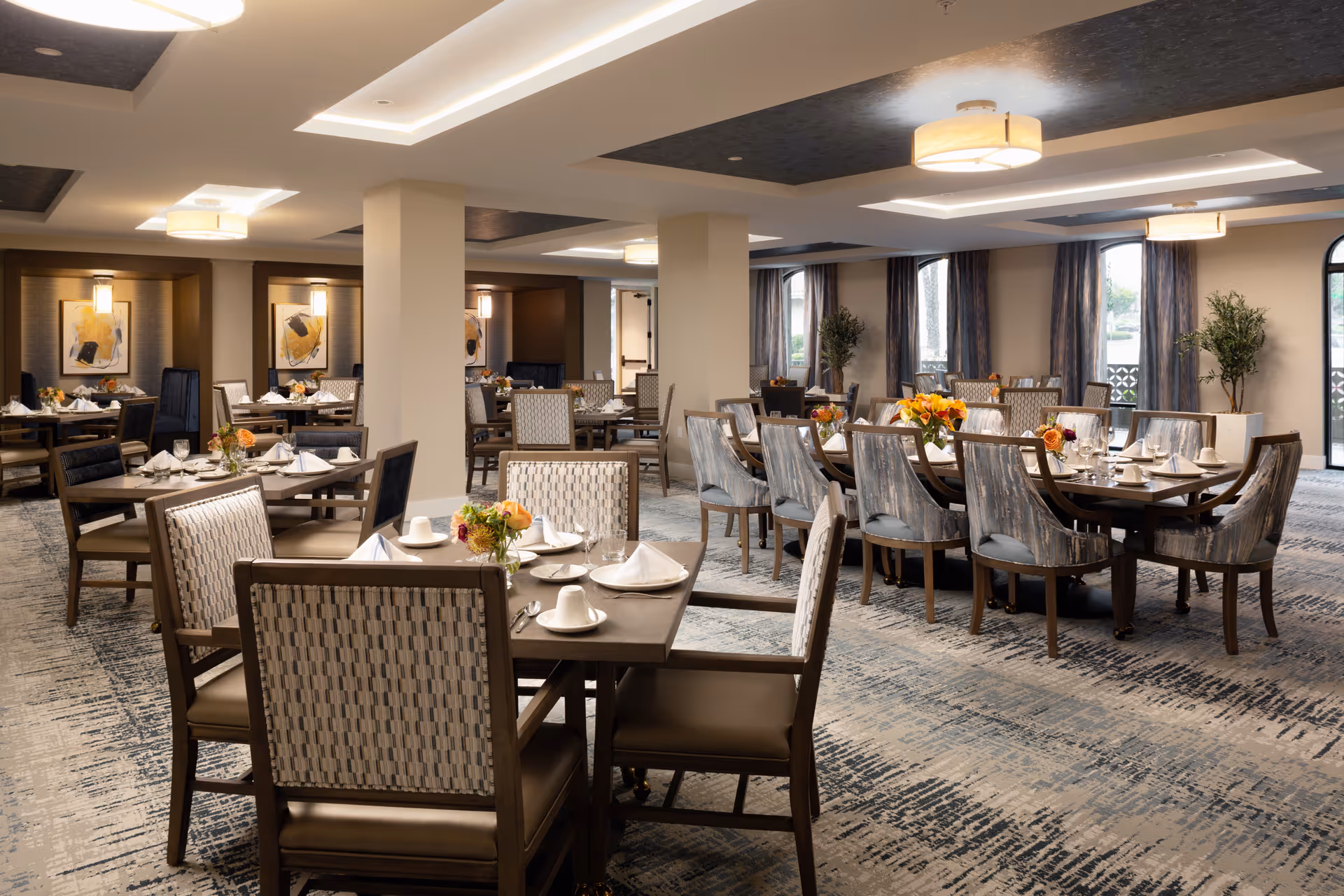 A spacious and well-lit dining room in a senior living facility with multiple tables set for a meal. Each table has chairs with patterned upholstery, white napkins, cups, plates, and floral centerpieces. The room features large windows with curtains, modern ceiling lights, and a patterned carpet.