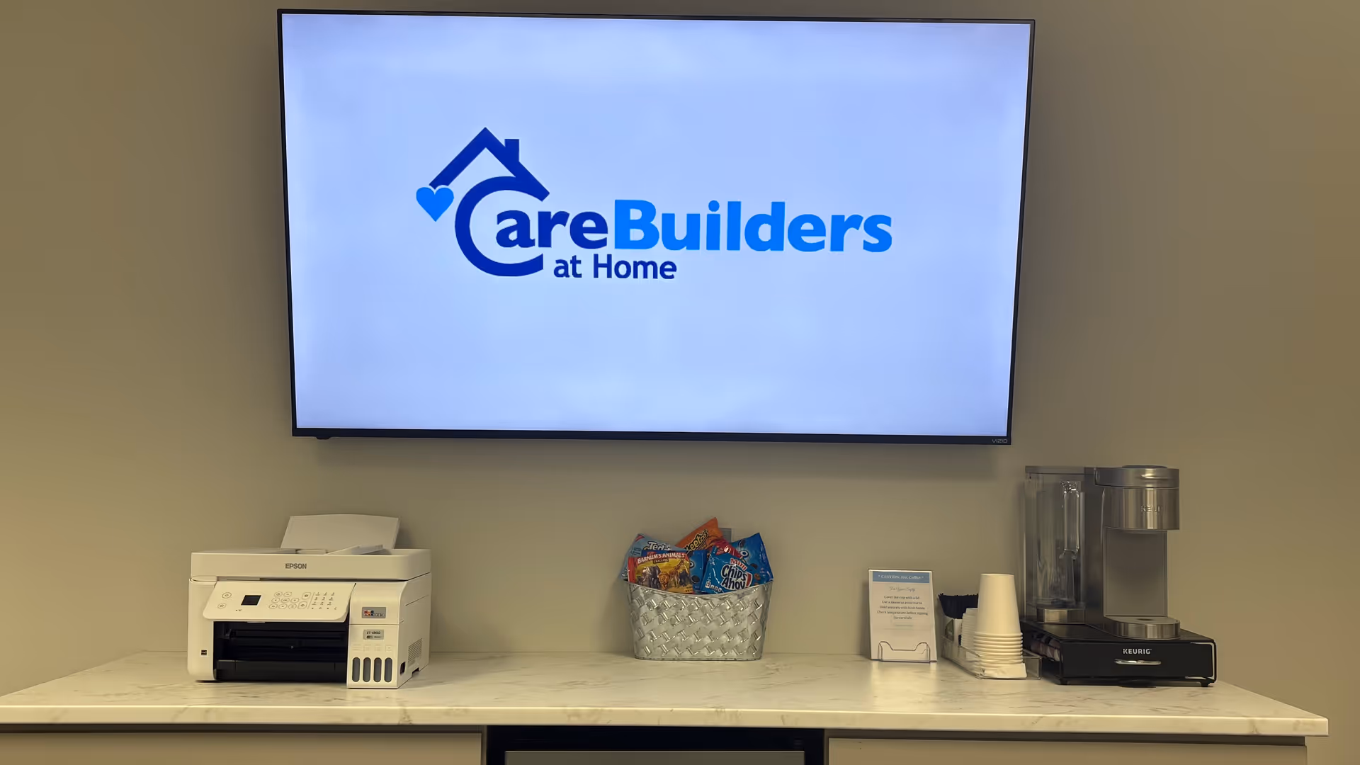 Large screen shows CareBuilders at Home logo; printer, snack basket, and coffee station on marble counter.