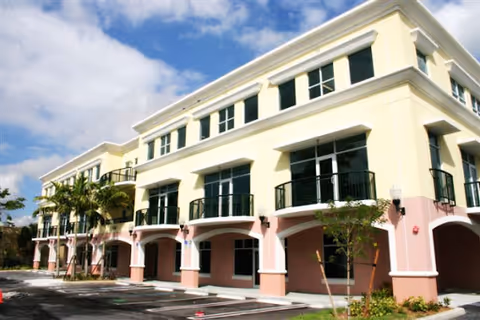 Pastel multi-story senior living facility exterior with arches, balconies, and palm trees.