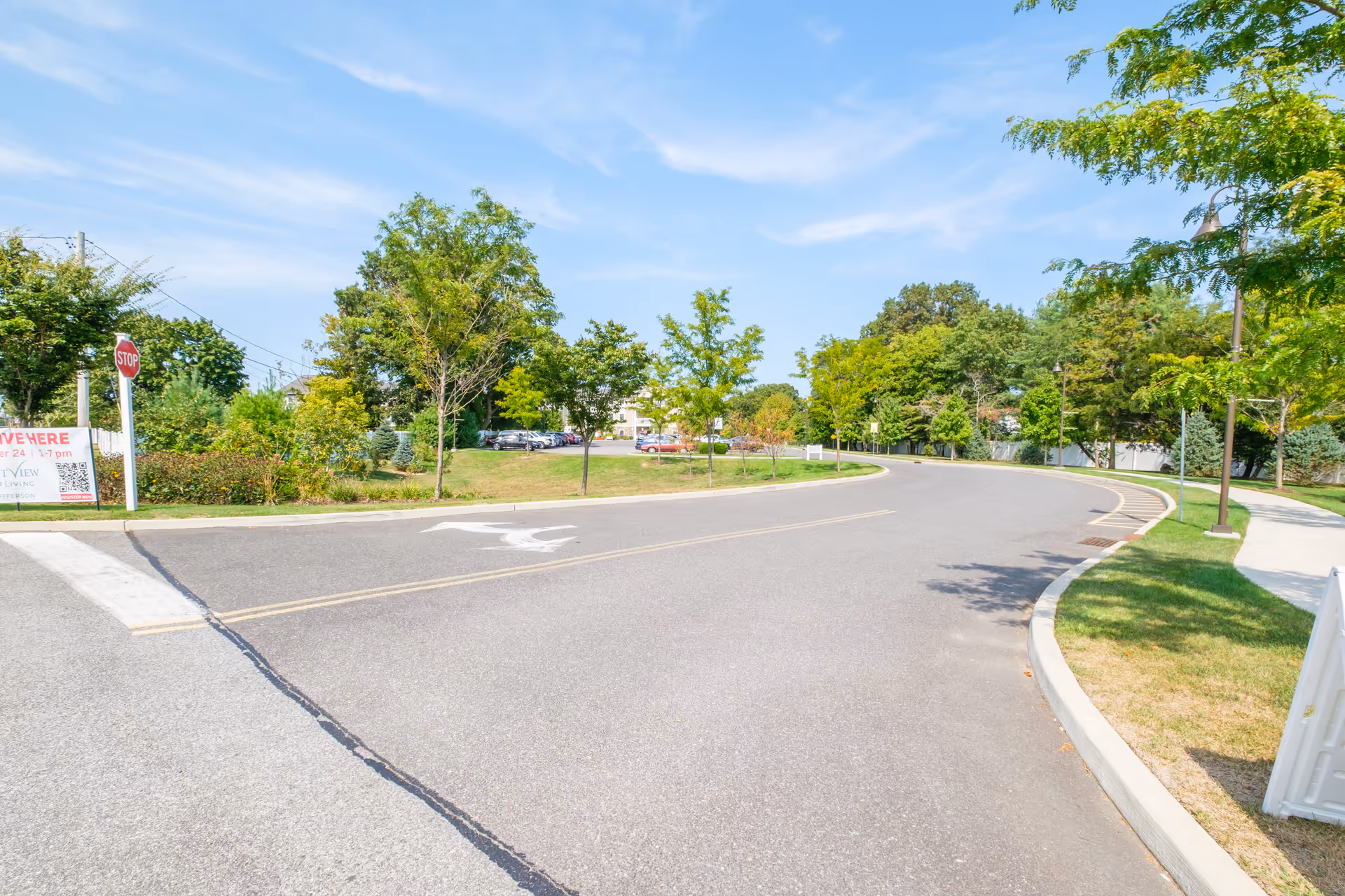 A curved road lined with trees and green grass on both sides under a blue sky with light clouds. There is a stop sign and a sign with some text on the left side near the road. A parking lot with several cars is visible in the background.