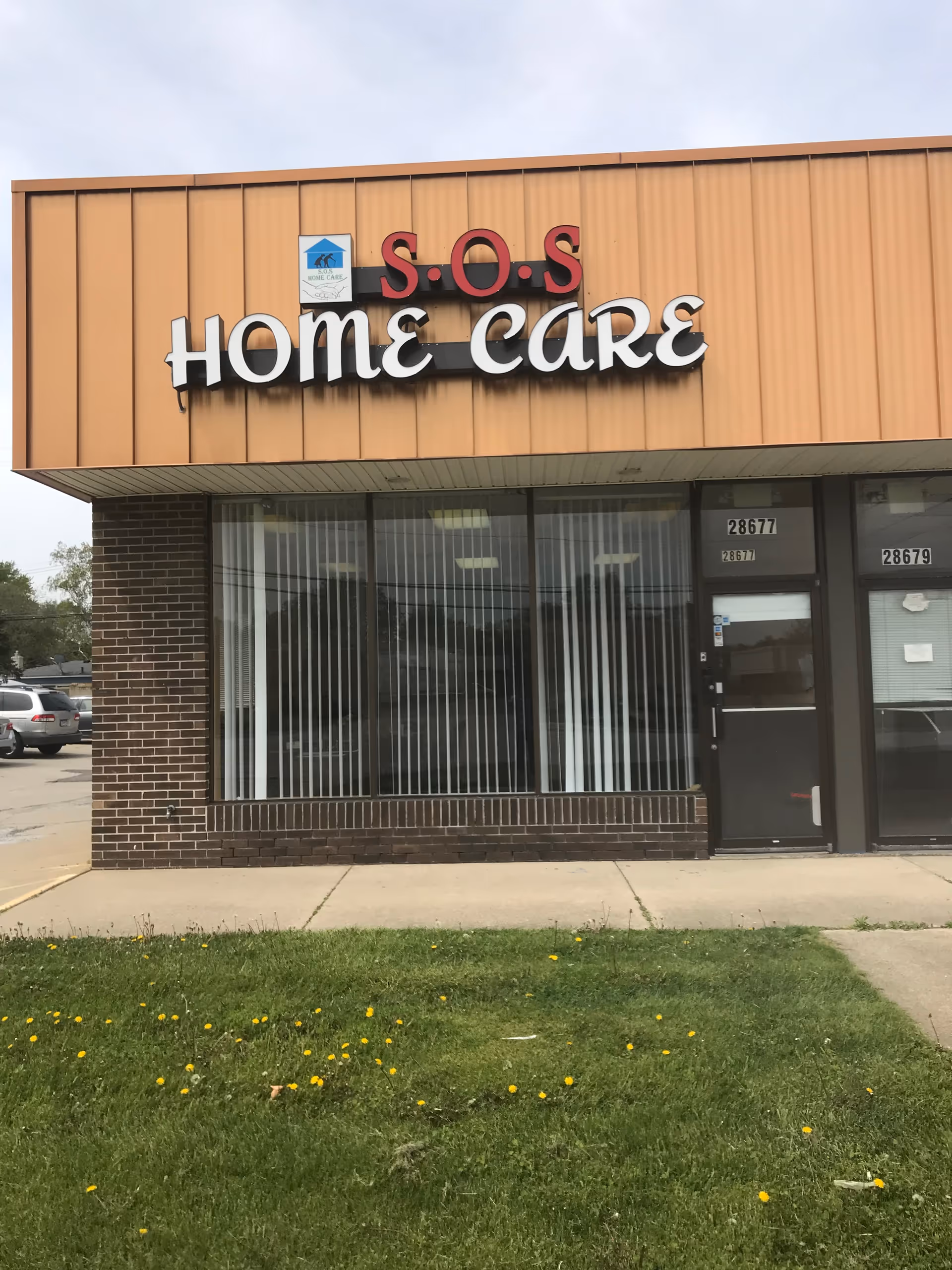 Orange storefront with SOS Home Care sign, large windows, brick base, and grassy foreground.