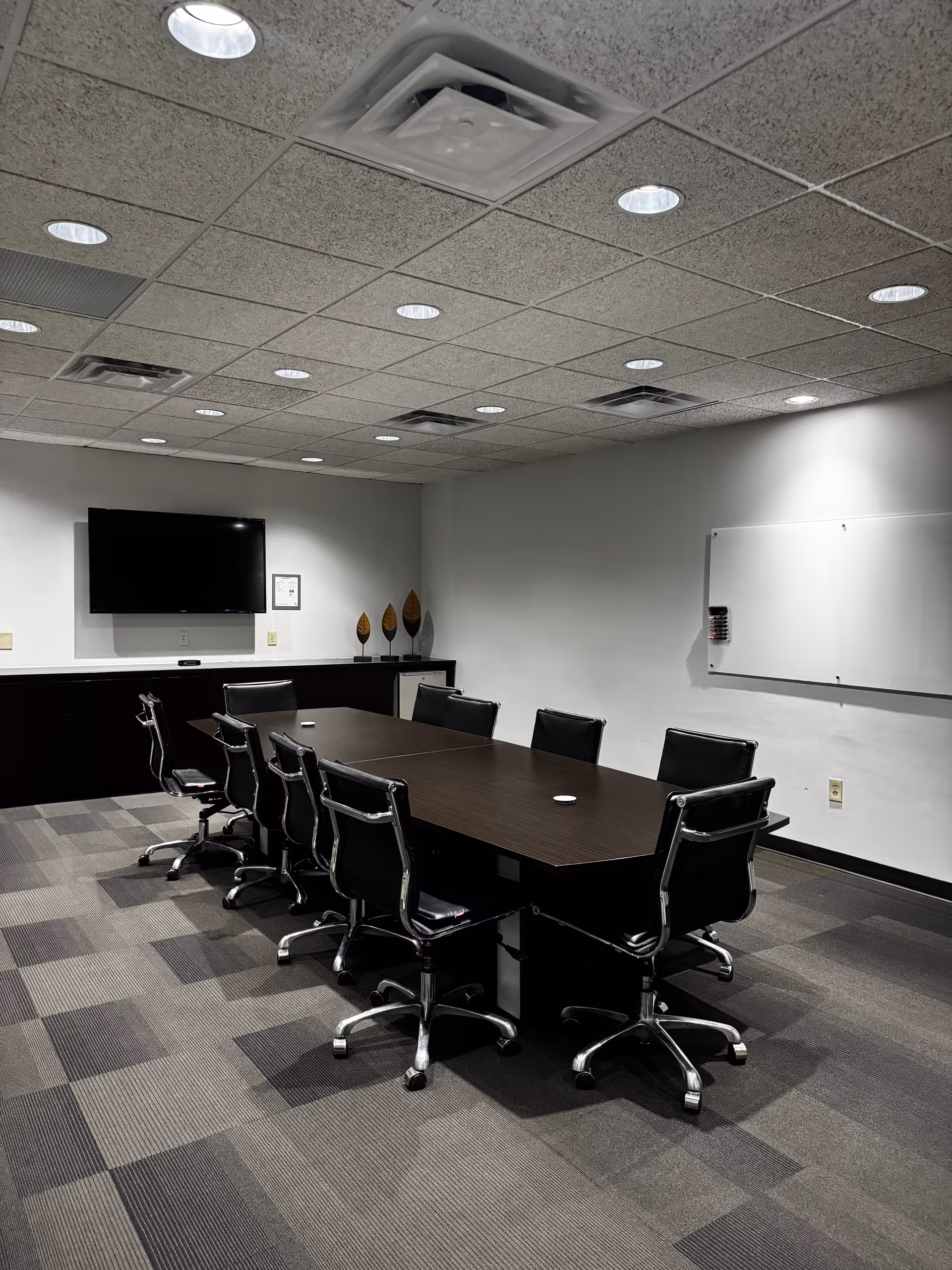 A modern conference room with a long dark table, black swivel chairs, TV, and whiteboard.
