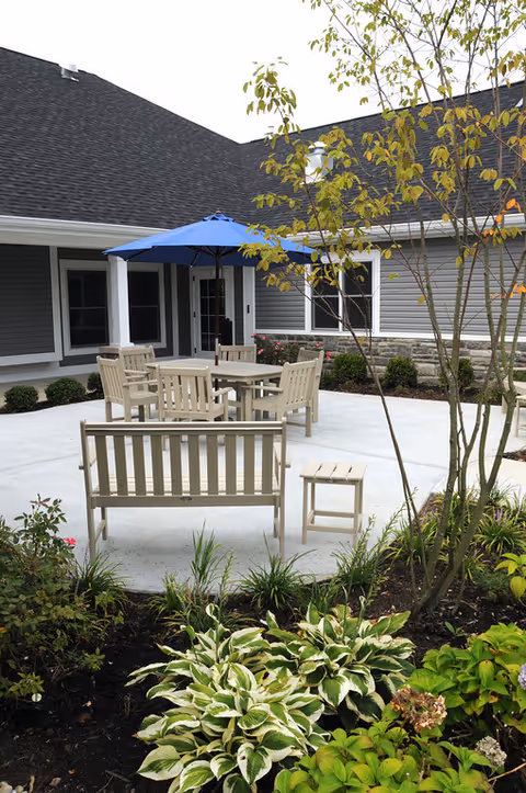 Outdoor patio area at Otterbein New Albany SeniorLife Neighborhood featuring beige wooden chairs and benches arranged around a table with a blue umbrella. The patio is surrounded by a garden with various green plants and small trees, and the building exterior is visible in the background with gray siding and white trim.