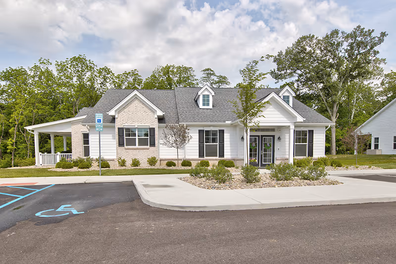 Single-story white senior living building with a covered porch, landscaped beds, and a handicap parking space in front.