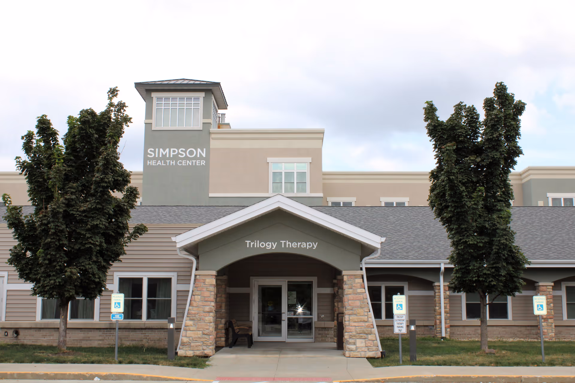 Front entrance of the Simpson Health Center building with a covered entry labeled 'Trilogy Therapy', flanked by trees and handicap parking signs.
