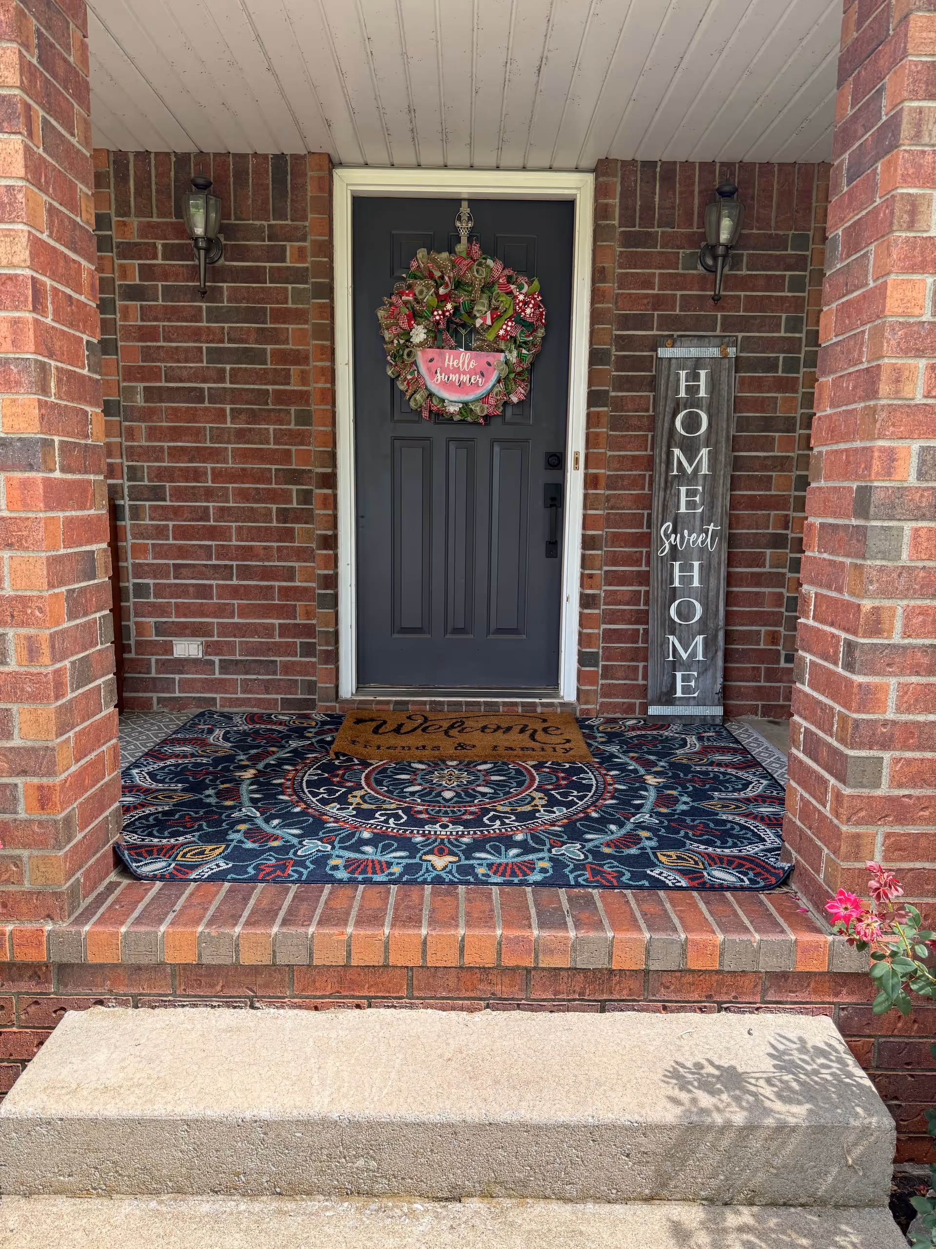 Brick front porch with a dark door, wreath, welcome mat, and vertical Home Sweet Home sign